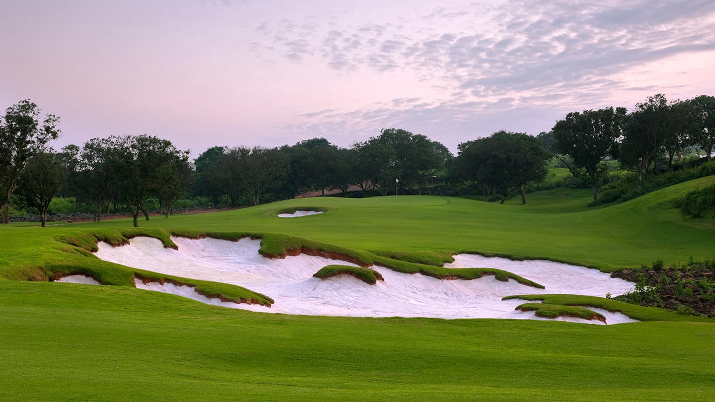 A green and sculpted white-sand bunker on the Blackstone Course at Mission Hills Haikou at dusk.