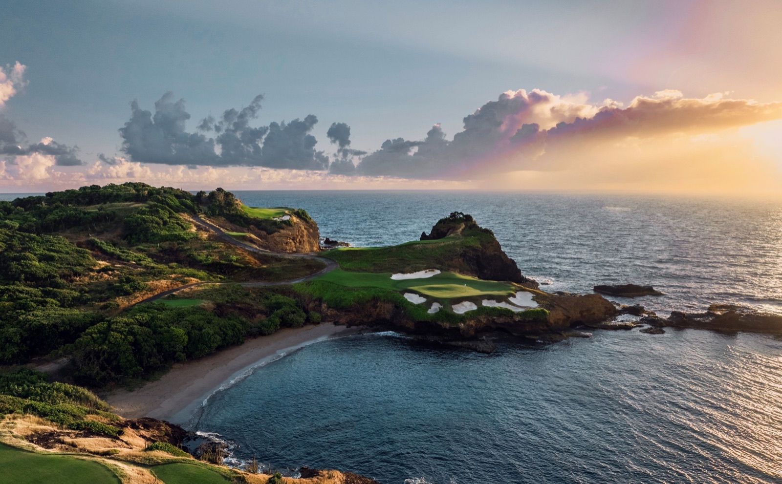 Elevated wide view of the 16th green at Point Hardy Golf Club, set on a rocky headland ringed by bunkers above a curling cove beach, with rock stacks in the surf and a pastel dusk sky.