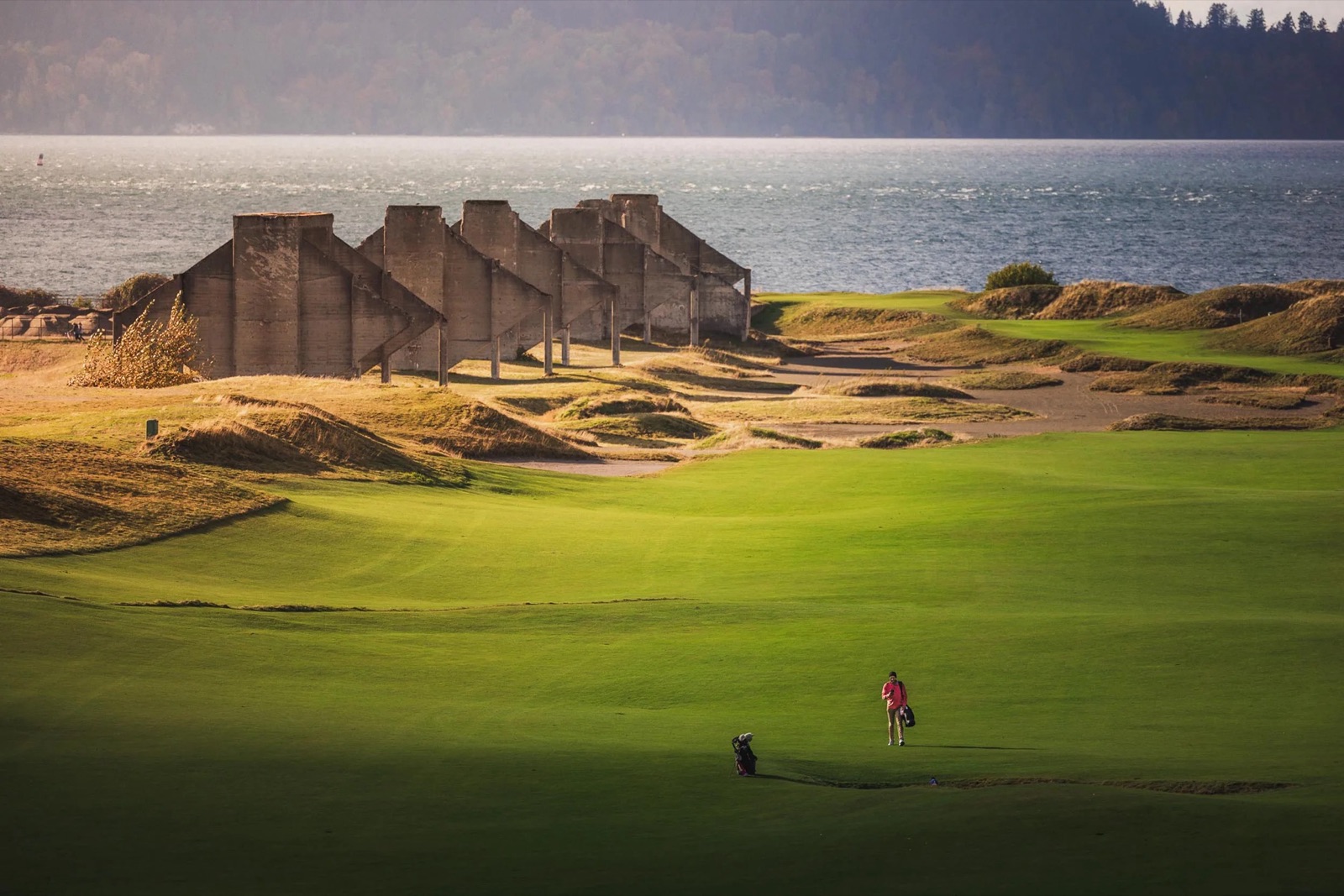 A golfer walking a fairway at Chambers Bay past the site's preserved concrete quarry ruins, Puget Sound beyond.