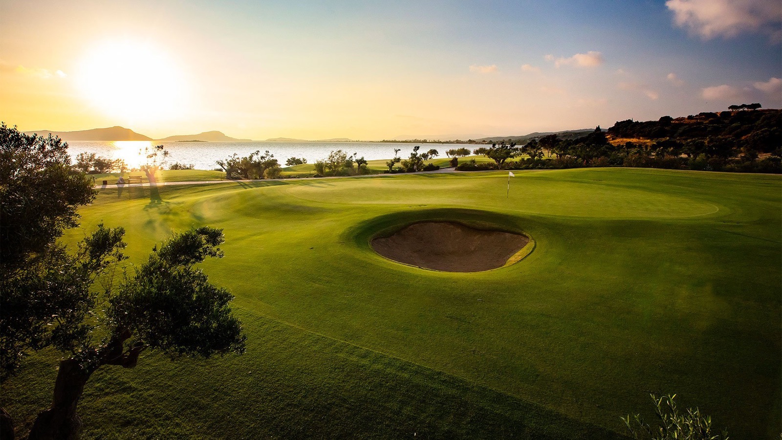 A green with flag on the Bay Course at Costa Navarino at golden hour, a deep round greenside bunker in front, the Bay of Navarino and its headlands directly behind and an olive branch framing the left of the frame.