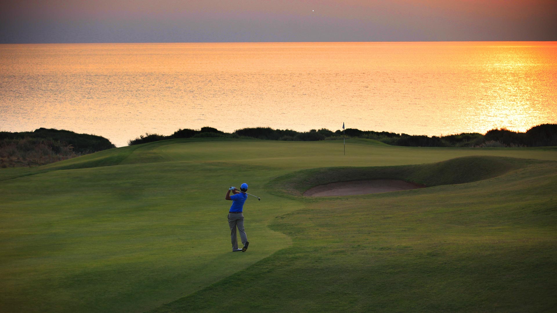 A golfer in blue at the top of his follow-through on the Dunes Course at Costa Navarino, playing to a green set above a gold-lit Ionian Sea with dune scrub between green and water and a pot bunker to the right.