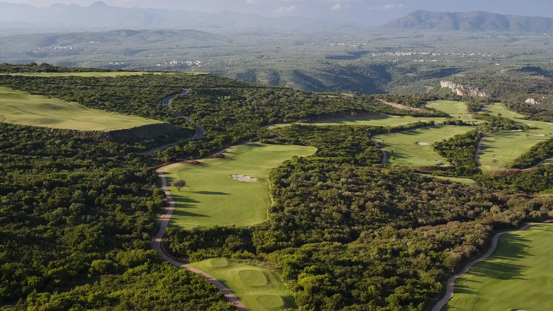 A wide elevated aerial across the Hills Course at Navarino Hills, several holes threading through dense maquis scrub with the olive-grove plain and the mountains of Messinia beyond, and no sea in view.