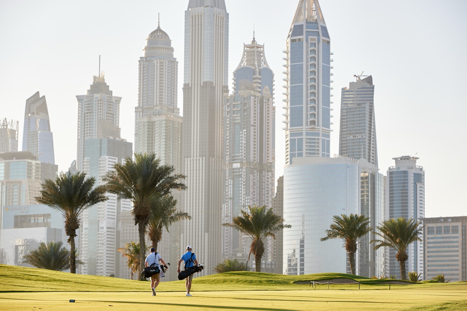 Two golfers walk a fairway on the Majlis Course at Emirates Golf Club, framed by the towers of Dubai Marina.