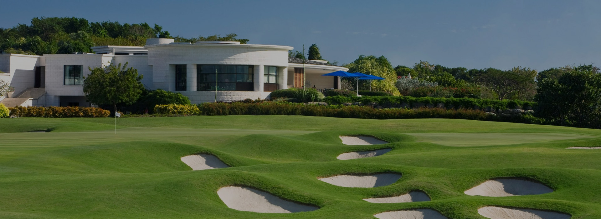 A green with a flag at The Country Club, Sandy Lane, fronted by a cluster of pot bunkers set in rolling ground, with the white clubhouse and blue parasols behind.