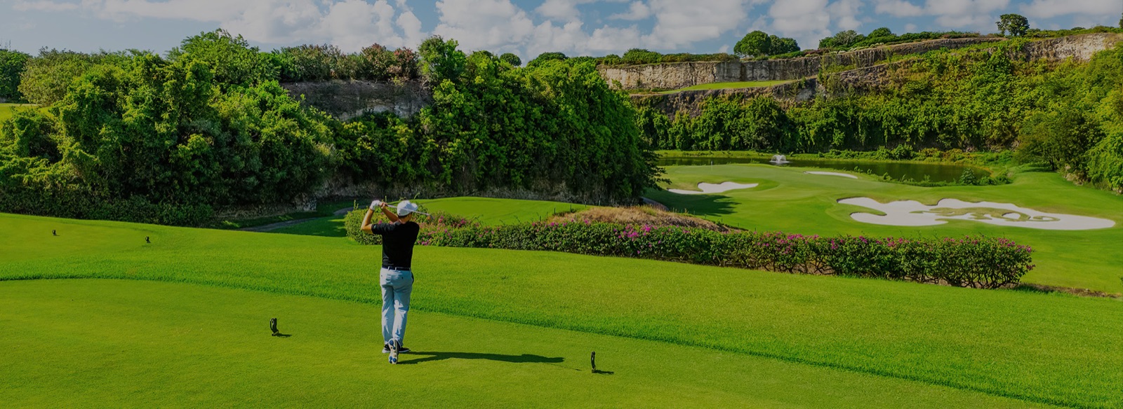 A golfer mid-swing on a wide fairway at The Green Monkey, Sandy Lane, with a vine-draped limestone quarry wall behind and a green beyond a water feature fronted by a large bunker complex shaped like a monkey.